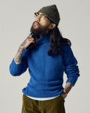 Male model wearing an Uskees ultra blue sweater and green beanie with tattoos on arms, standing against a plain background