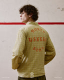 Back view of a man wearing a yellow checkered Uskees jacket with text, standing against a textured wall.