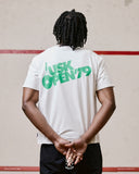 Back view of male model wearing an Uskees white short-sleeve t-shirt with green text on the back, standing in front of a wall.