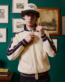 Person wearing a cream-colored jacket with dark sleeves and a cap, standing against a wall with framed pictures.