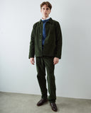 A male model wearing a dark green corduroy shirt-jacket and matching corduroy trousers, styled with a blue knit sweater, white collared shirt, and dark brown leather loafers, against a plain light gray wall and concrete floor.