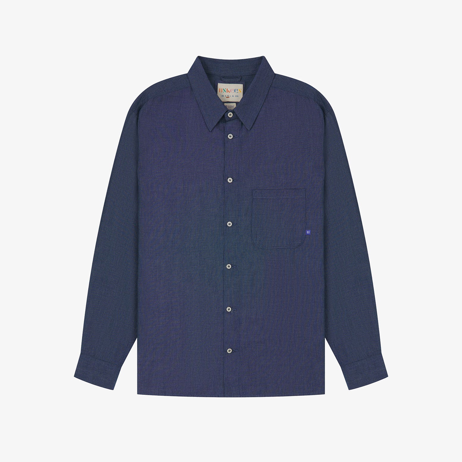 Flat front view of mid blue organic chambray Oxford shirt from Uskees. Clearly showing loose fit and 8-button front.