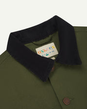 Close-up view of uskees cotton canvas chore jacket for men in coriander green showing the dark blue cord collar, brand label and metal buttons 