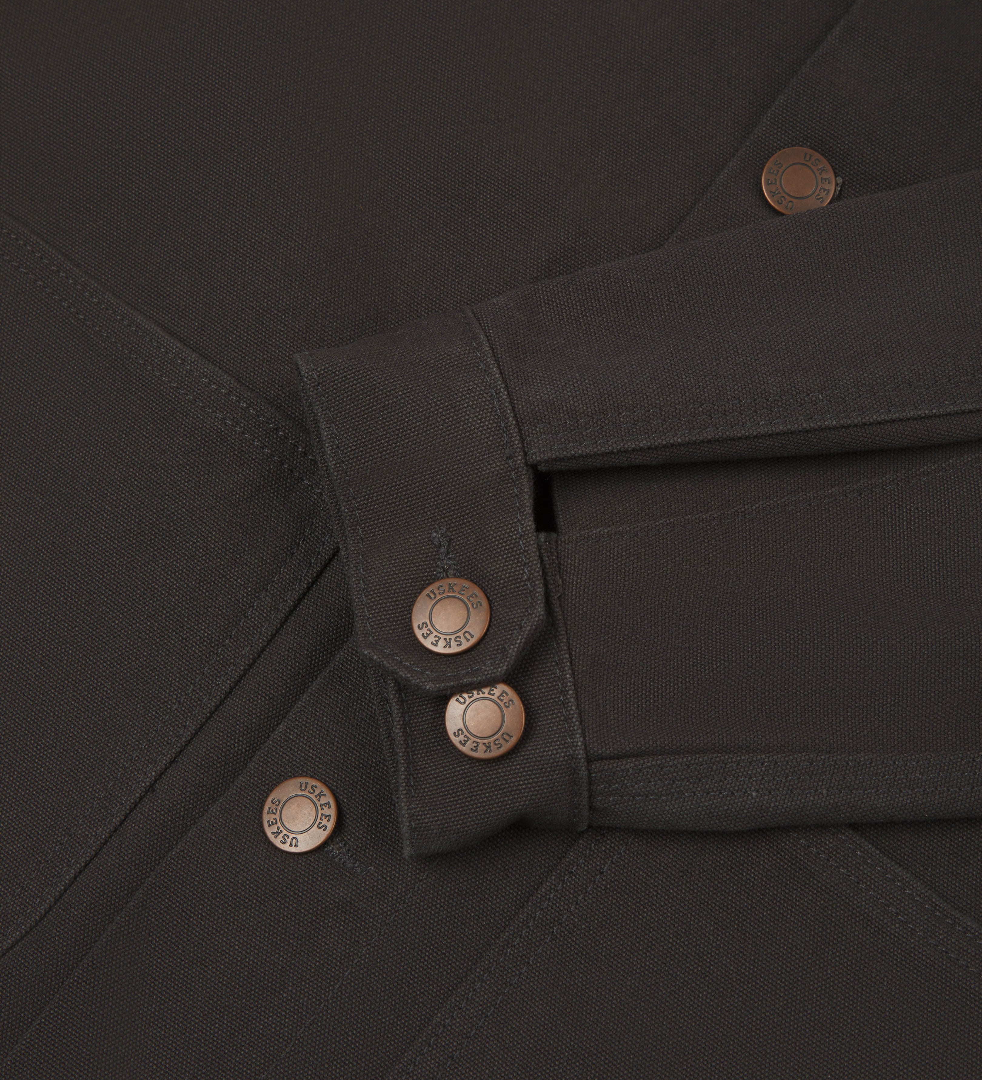 Detail shot of uskees  men's canvas chore jacket in charcoal with a green collar showing the adjustable cuff, sleeve and pockets in close-up 