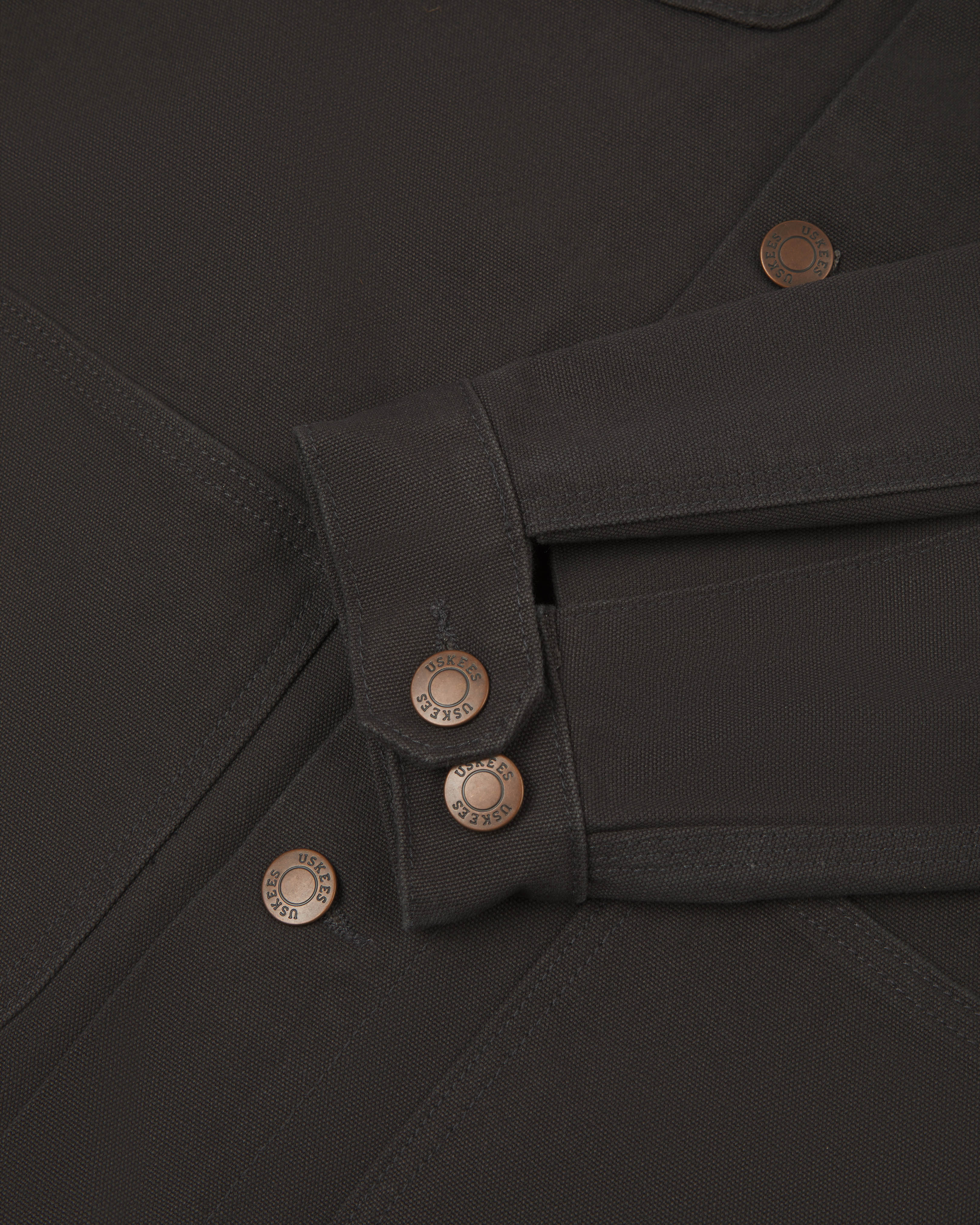 Detail shot of uskees  men's canvas chore jacket in charcoal with a green collar showing the adjustable cuff, sleeve and pockets in close-up 