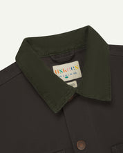 Front close-up shot of uskees cotton canvas chore jacket for men in dark grey. Clear view of the green corduroy collar, brand label and buttons