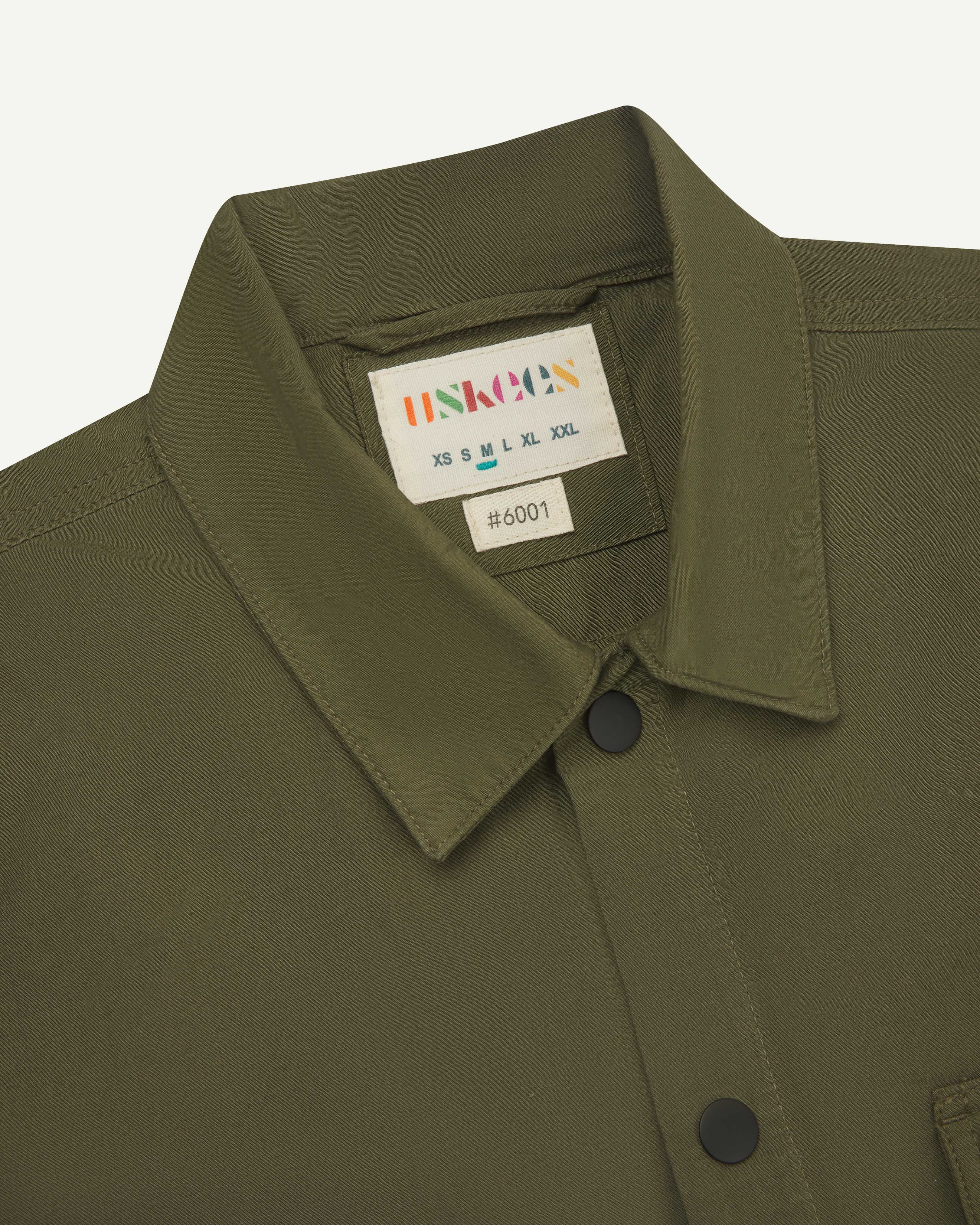 front close shot of men's olive green uskees lightweight shirt with black popper front fastening. Clear view of collar and brand/size label at neck.
