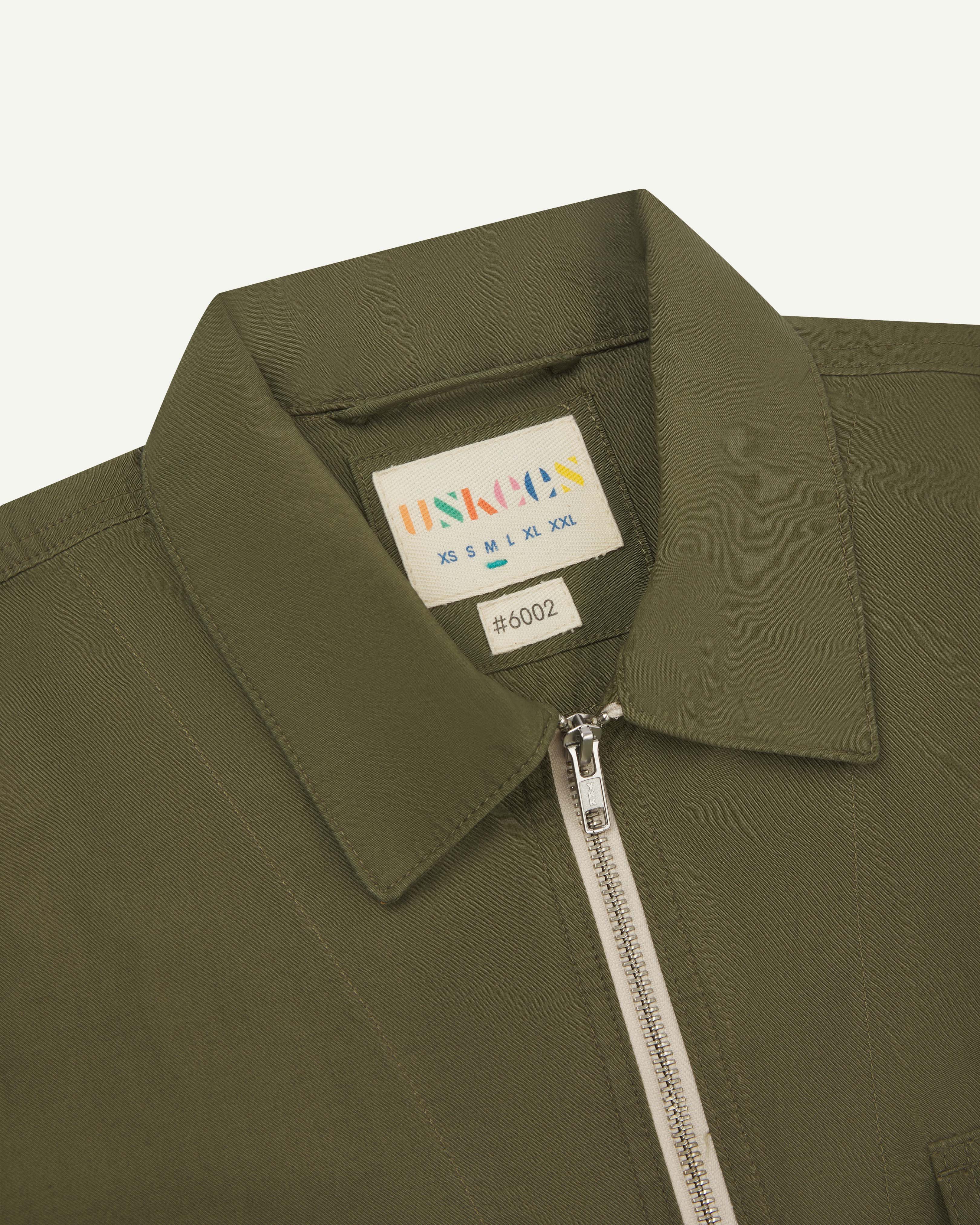 Close-up shot of uskees zip front lightweight men's jacket in olive green showing the contrast front zip and brand/size label.