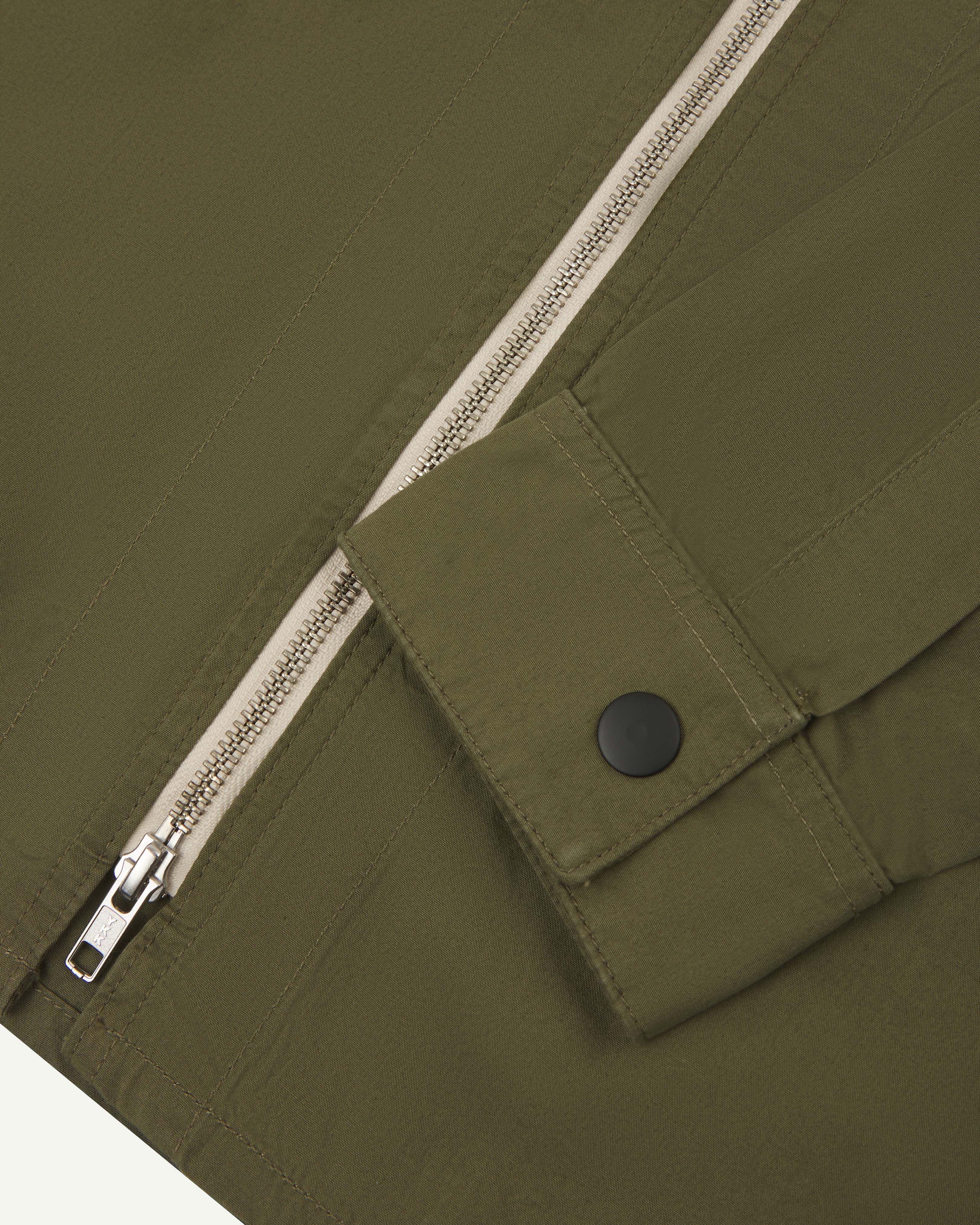 Close-up view of uskees zip front lightweight men's jacket in olive green showing the contrast zip and black popper fastening on cuff.