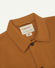 Close-up front flat shot of uskees lightweight tan men's shirt with short sleeves showing collar, uskees neck label & black popper fastenings