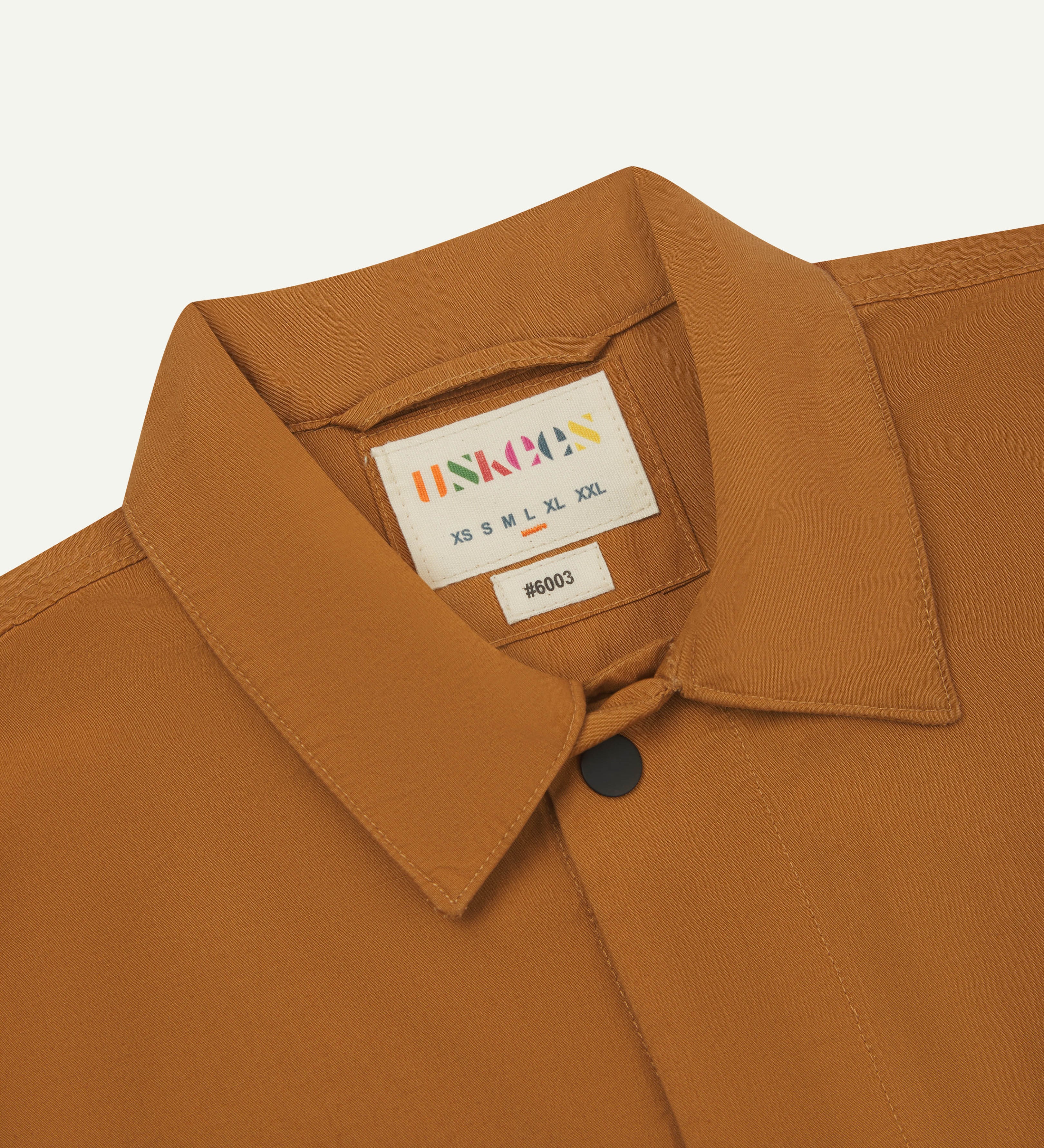 Close-up front flat shot of uskees lightweight tan men's shirt with short sleeves showing collar, uskees neck label & black popper fastenings