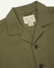 Front close view of moss green organic cotton drill commuter blazer with patch pockets showing collar, corozo buttons and Uskees branding label.