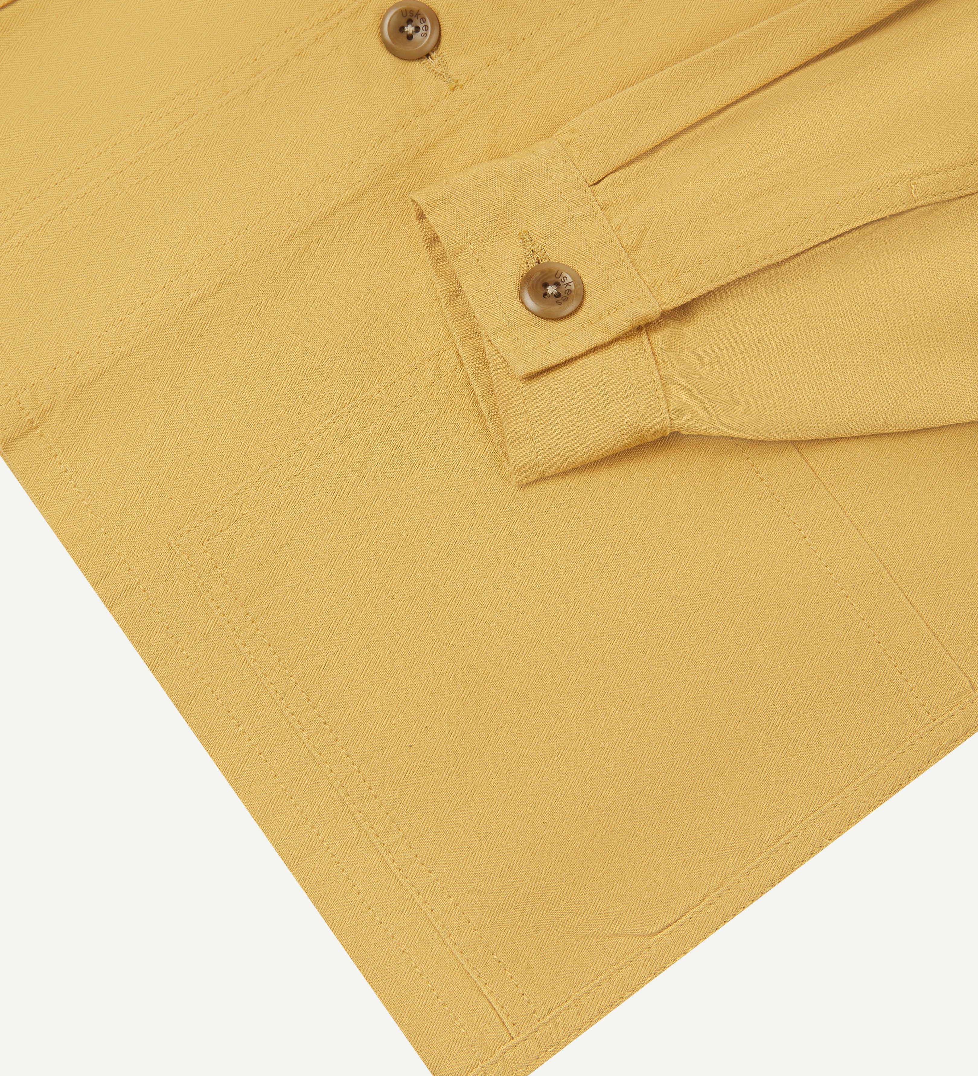 Close-up view of one of two hip pockets, cuff and Uskees branding tag of the 3041 signal jacket. Also is a closer view of the herringbone texture of the sandstone-yellow cotton.