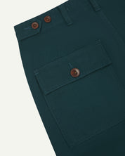 Close-up angled view of Uskees peacock coloured cotton work pants with focus on natural corozo buttons, left rear pocket, belt loops, triple stitching and adjustable button waist.