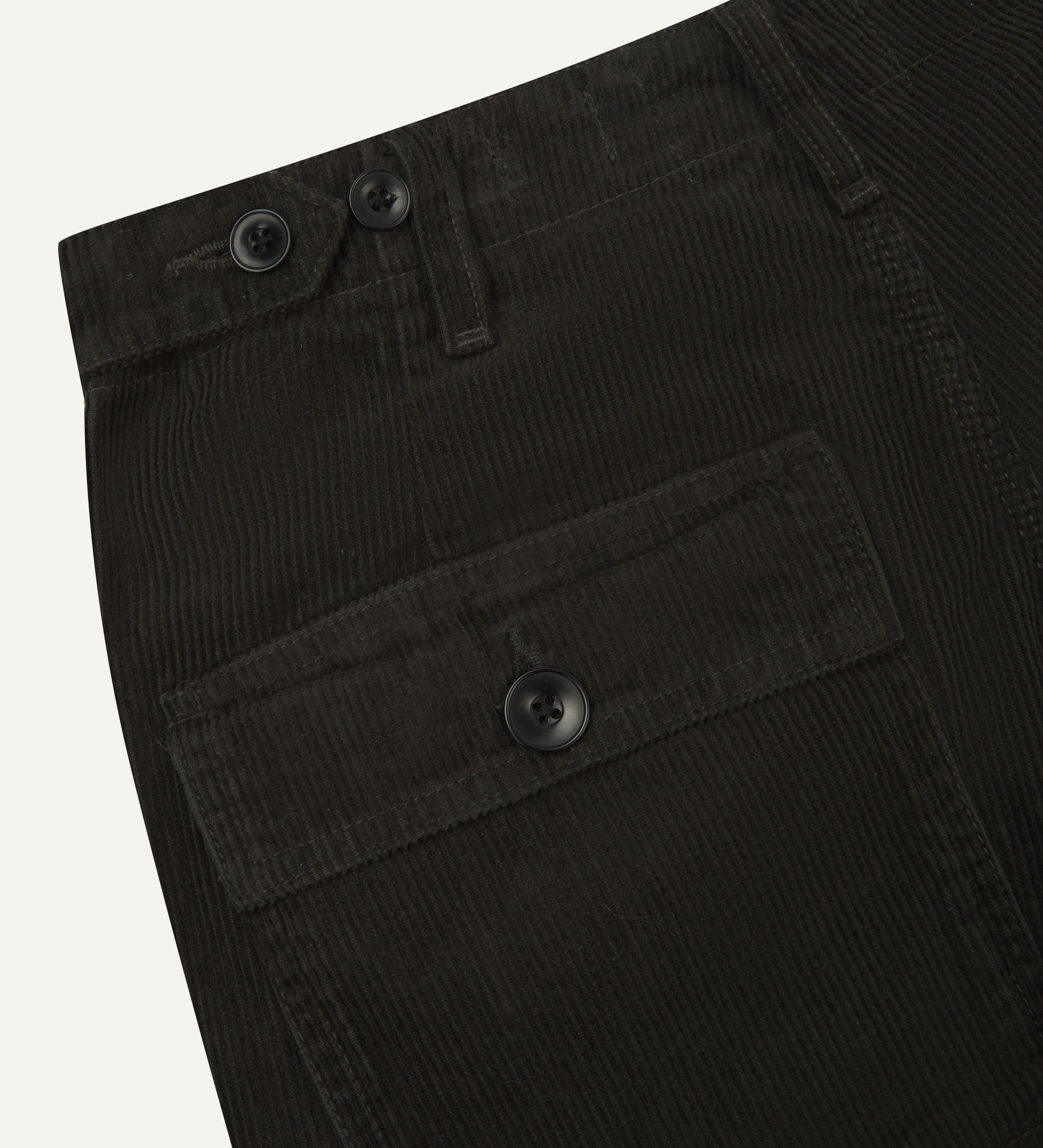 Angled, close-up view of Uskees faded-black corduroy work pants with focus on natural corozo buttons, left rear pocket, belt loops, triple stitching and adjustable button waist.