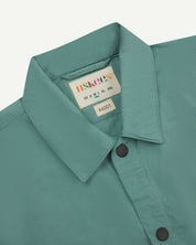 front flat shot of eucalyptus green uskees lightweight shirt with popper front fastening. Shows collar and brand/size label in close-up.