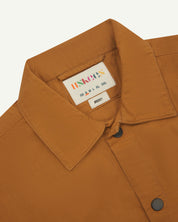 front flat shot of light brown uskees lightweight shirt with popper front fastening. Shows collar and brand/size label in close-up.