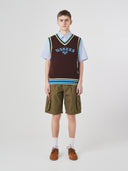 Front shot of male model wearing a brown sleeveless sweater with 'USKEES' branding, light blue shirt, green cargo shorts, and brown shoes on a white background.