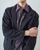 A male model wearing a black utility jacket over a blue plaid button-down shirt, shown from the chin down to the waist against a light gray studio background.