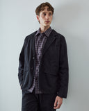Male model standing in a studio wearing a black utility-style blazer over a blue plaid button-front shirt, paired with matching trousers.