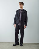 Male model standing in a studio wearing a black utility-style jacket over a blue plaid button-down shirt, paired with matching trousers and black leather shoes.