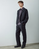 Male model standing in a studio wearing an Uskees black utility-style jacket over a blue plaid button-down shirt, paired with matching trousers and black leather shoes.
