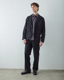 A male model wearing Uskees black utility blazer, black trousers, a blue checkered shirt, and black leather shoes, standing against a light gray wall.