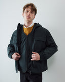 A man wearing an Uskees dark grey hooded zip-up coat over a liner jacket and mustard yellow sweater and white shirt, standing against a plain light gray wall.