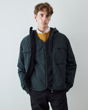 A man wearing an Uskees dark grey hooded zip-up coat over a liner jacket, mustard yellow sweater and white collared shirt, standing against a light gray studio background.