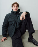 A man wearing an Uskees dark grey hooded utility jacket, dark blue trousers, black socks, and black leather loafers sits against a light gray wall on a concrete floor.
