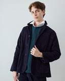 A man wearing an Uskees dark navy corduroy utility jacket over a green patterned sweater and white collared shirt, photographed against a light gray studio background.