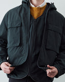 A close-up of a person wearing a charcoal hooded coat with multiple flap pockets, layered over an Uskees evo liner jacket and mustard yellow knit sweater and white collared shirt.