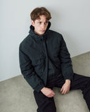 A man wearing an Uskees dark charcoal gray hooded coat and dark blue pants, sitting on a concrete floor against a light gray wall.