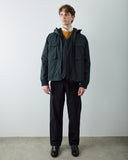 A man wearing an Uskees dark charcoal hooded utility jacket over a mustard yellow sweater, black trousers and black shoes, standing against a plain light gray wall.