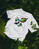 Back view of an Uskees white short-sleeve t-shirt featuring a detailed graphic of a flying bird with vibrant green, blue, and black wings, laid flat on a bed of green ferns and foliage.