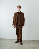 A male model stands in a minimalist studio wearing an Uskees brown corduroy shirt jacket and matching brown corduroy trousers, layered over a dark red sweater, white collared shirt, and dark brown leather shoes.