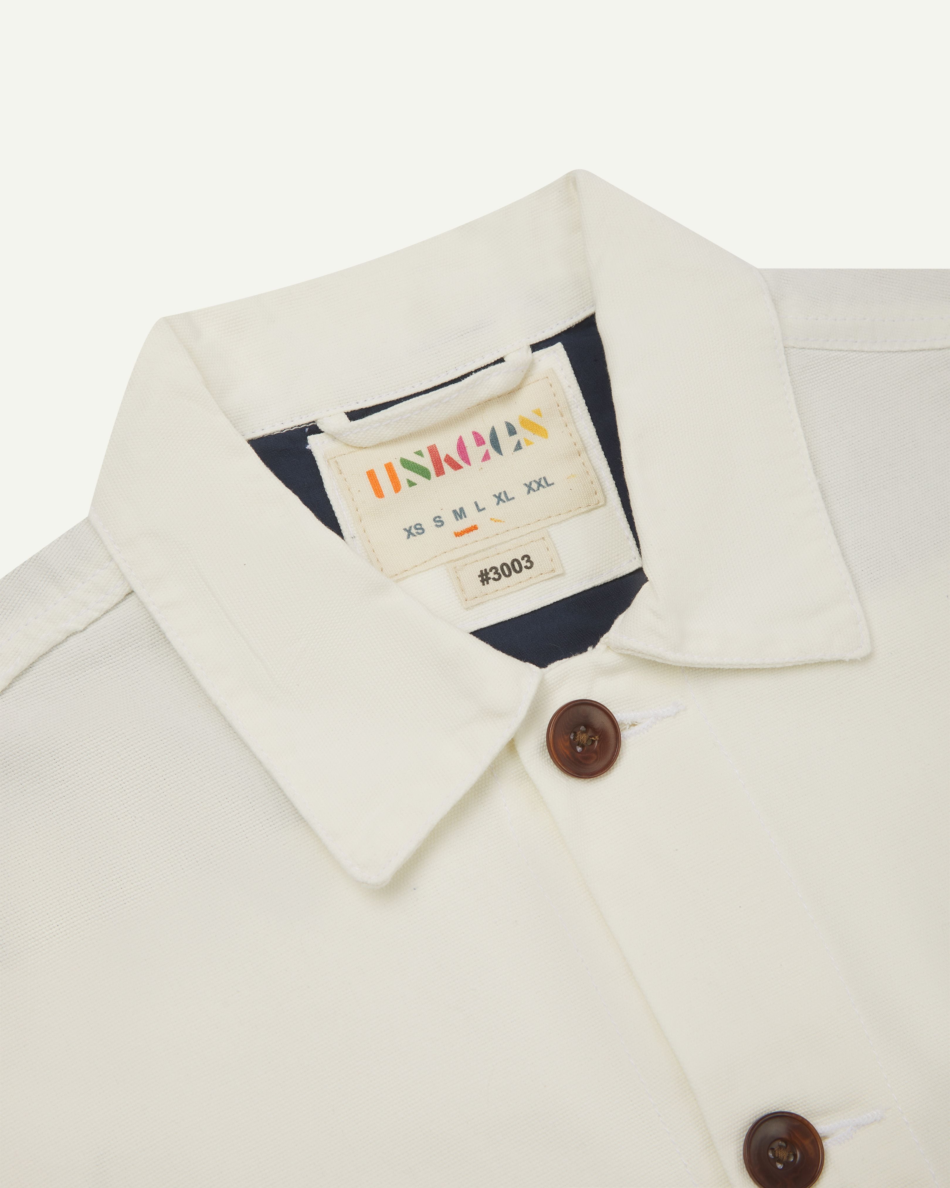 Front close-up view of cream buttoned 3003 workshirt from Uskees. Showing brand label, navy yoke lining and brown corozo buttons