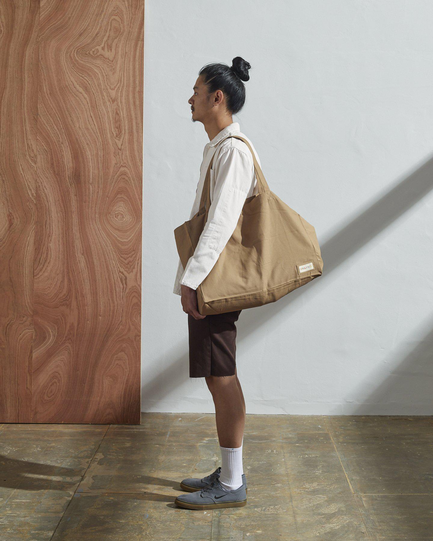 Full-length, front-view of model carrying Uskees #4001, large khaki tote bag over left shoulder. Paired with #3002, light grey zip-front jacket.