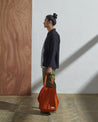 Full-length, side-view of model holding Uskees #4002, small golden-orange tote bag.