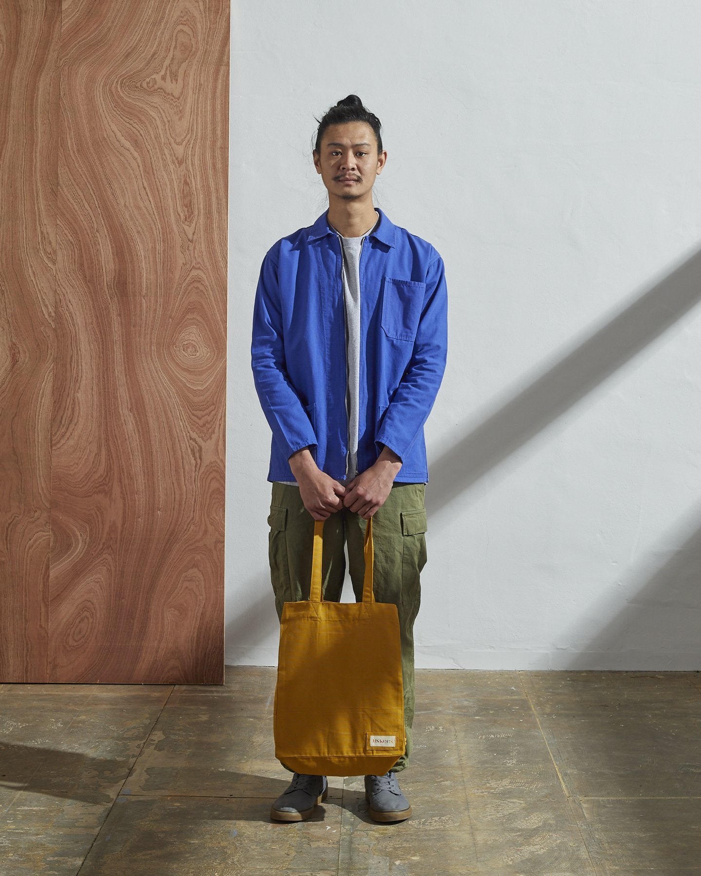 Full-length, front-view of model carrying Uskees #4002, small yellow tote bag. Paired with #3002, blue zip-front jacket.