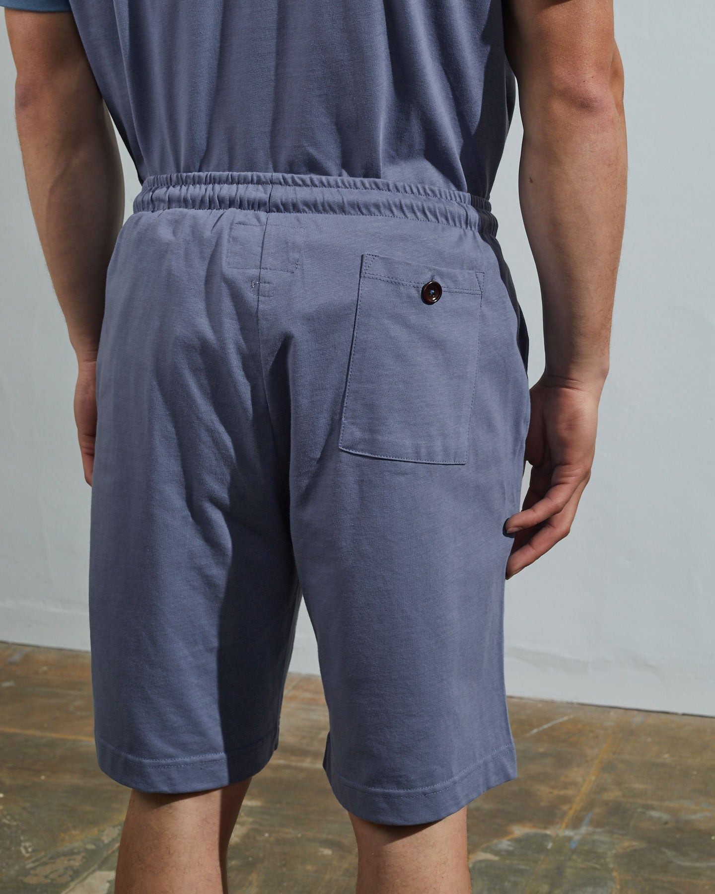 Back view of model wearing teal, relaxed cut organic cotton #7007 jersey shorts by Uskees with focus on back pocket with corozo button.