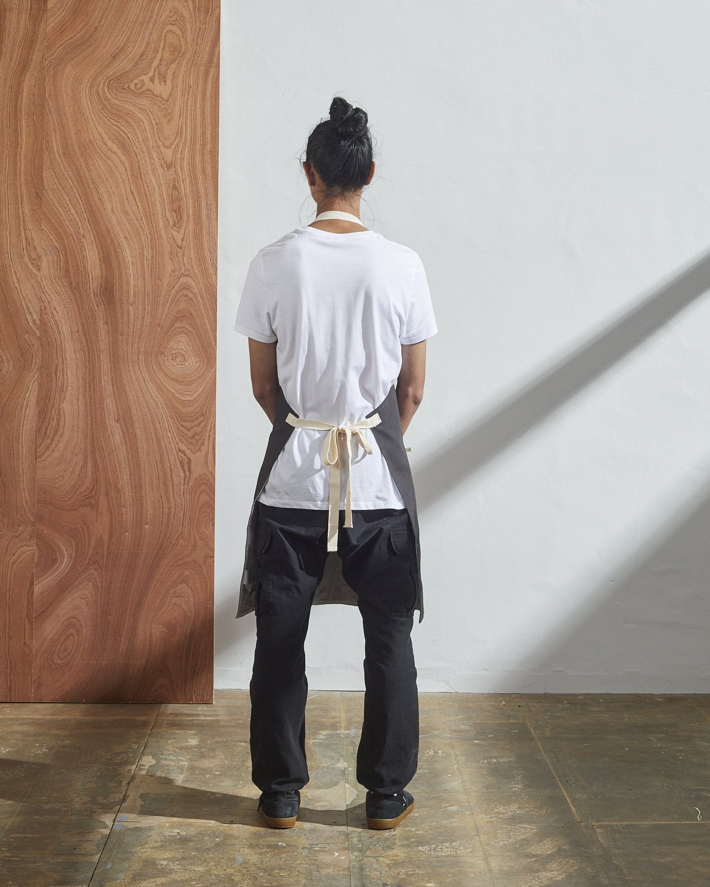 Full-length, rear view of model wearing Uskees #9004 faded-black work apron by Uskees. Clear view of cream rear ties.