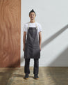 Full-length, front view of male model wearing Uskees #9004 faded-black carpenter-style apron by Uskees. Showing all three pockets and contrasting cream straps.
